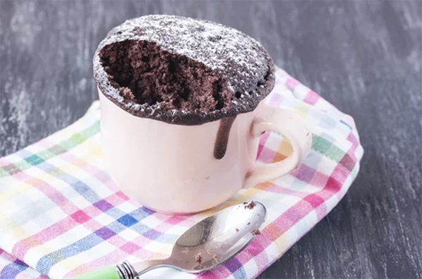 Κέικ σε κούπα πασπαλισμένο με άχνη ζάχαρη, επάνω σε μια καρό, πολύχρωμη πετσέτα.